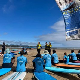 Surf instructors in yellow shirts teach students in blue gear on a sandy beach with surfboards.