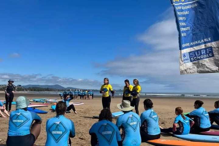 Surf instructors in yellow shirts teach students in blue gear on a sandy beach with surfboards.