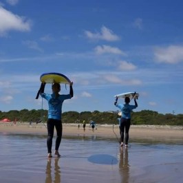 Two people in blue shirts holding surfboards on a sunny beach.