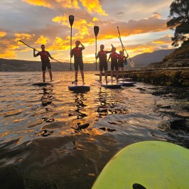 Four people paddleboarding on a lake at sunset, with an orange sky and distant hills.