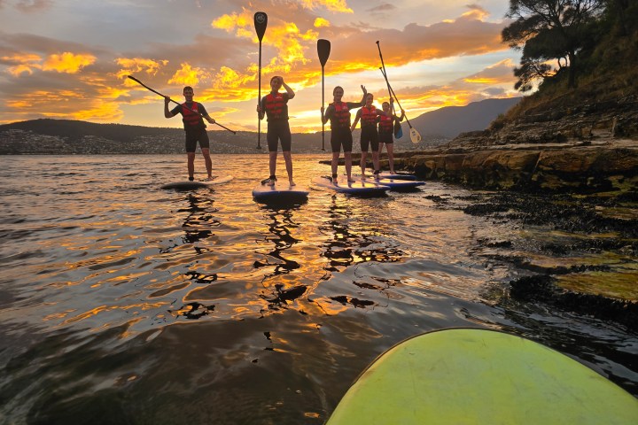 Four people paddleboarding on a lake at sunset, with an orange sky and distant hills.