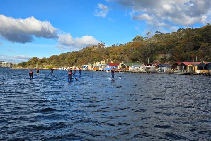 People paddleboarding on a river with colorful boathouses and trees in the background under a cloudy sky.