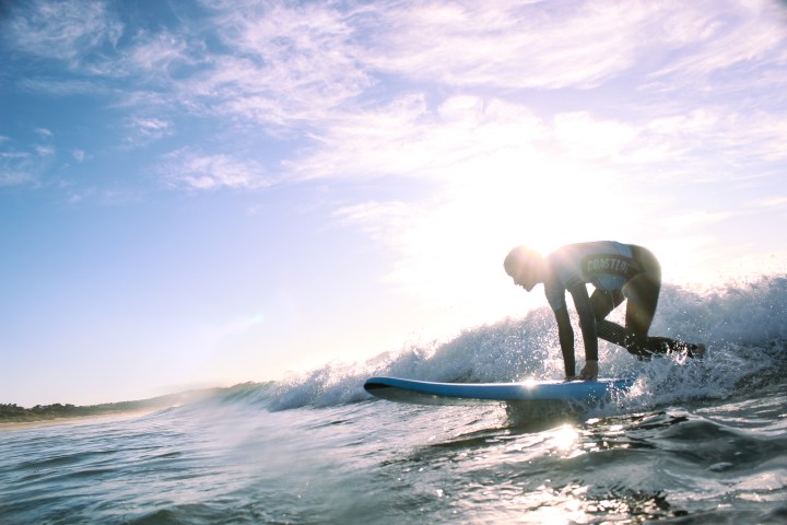Person surfing on a wave at sunrise, with a bright sun and blue sky in the background.