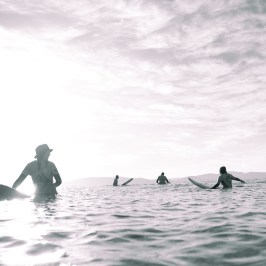 Silhouettes of surfers with boards in the ocean under a bright, cloudy sky.