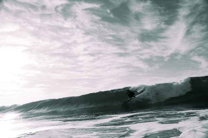 Surfer riding a large wave under a cloudy sky in black and white.