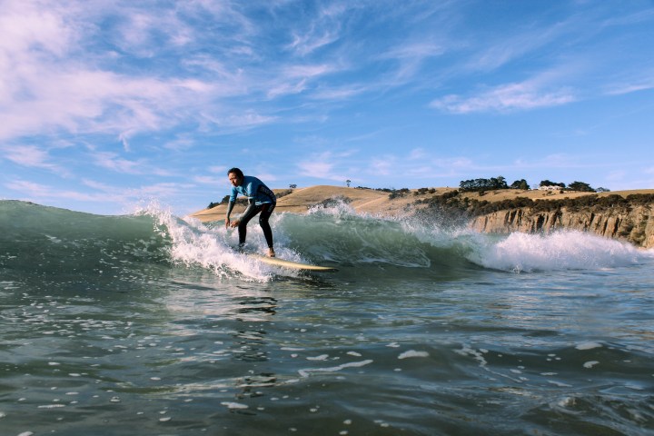 Surfer riding a wave near a rocky coastline under a clear blue sky.