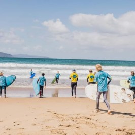 Group of surfers in blue and yellow shirts entering the ocean on a sunny day.