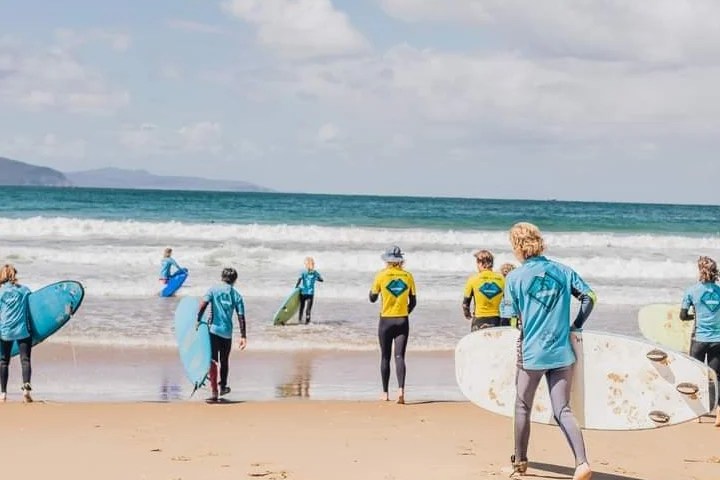 Group of surfers in blue and yellow shirts entering the ocean on a sunny day.