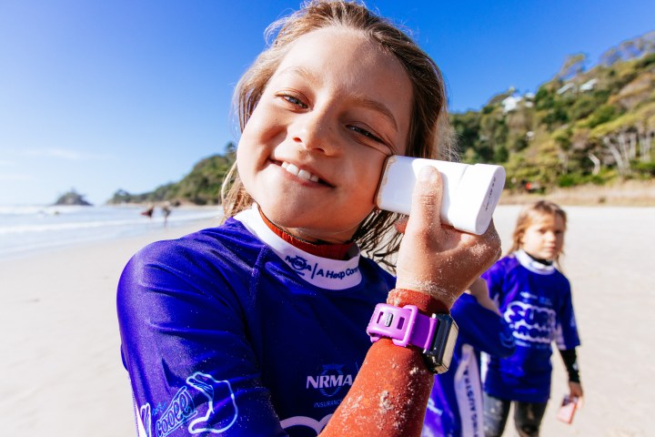 Young girl in purple wetsuit applying sunscreen at a sunny beach.