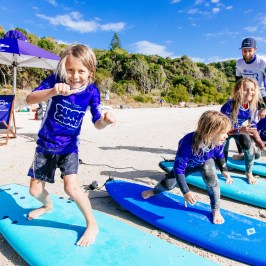 Kids practice surfing on the beach under a coach's guidance, wearing blue shirts and standing on surfboards.