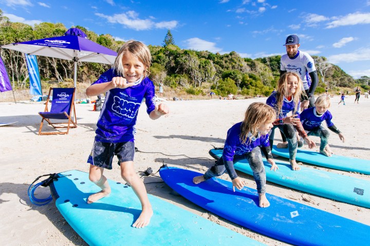 Kids practice surfing on the beach under a coach's guidance, wearing blue shirts and standing on surfboards.
