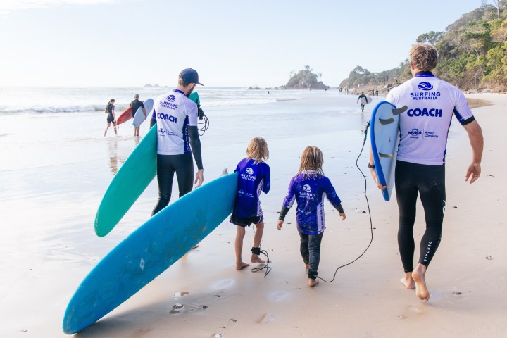 Surfers on a beach, two adults with boards and two children, walking towards the ocean.