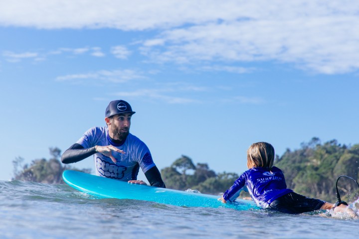 Adult and child surfing on a blue surfboard in the ocean, with trees in the background.