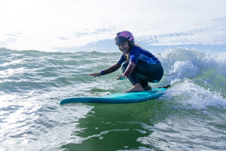 Young surfer with helmet riding a wave on a blue surfboard with a joyful expression.